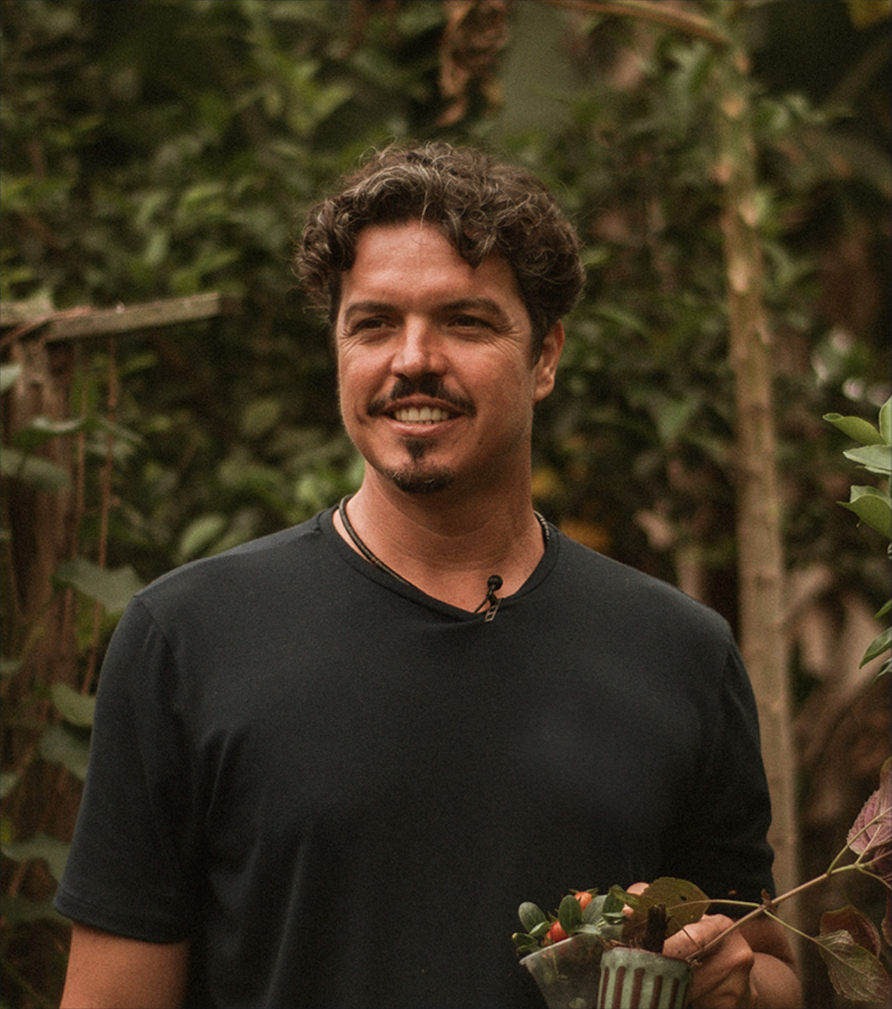 Marquito de blusa preta com algumas plantas em sua mão esquerdo olhando levemente para seu lado direito e sorrindo
